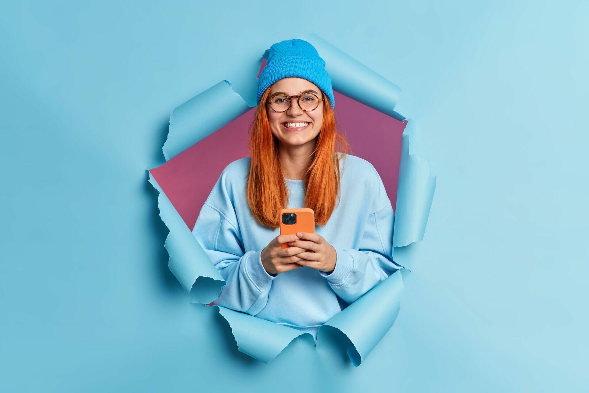 happy-smiling-millennial-girl-with-red-hair-holds-modern-cellular-enjoys-texting-in-social-media-uses-mobile-network-services-wears-blue-jumper-and-hat