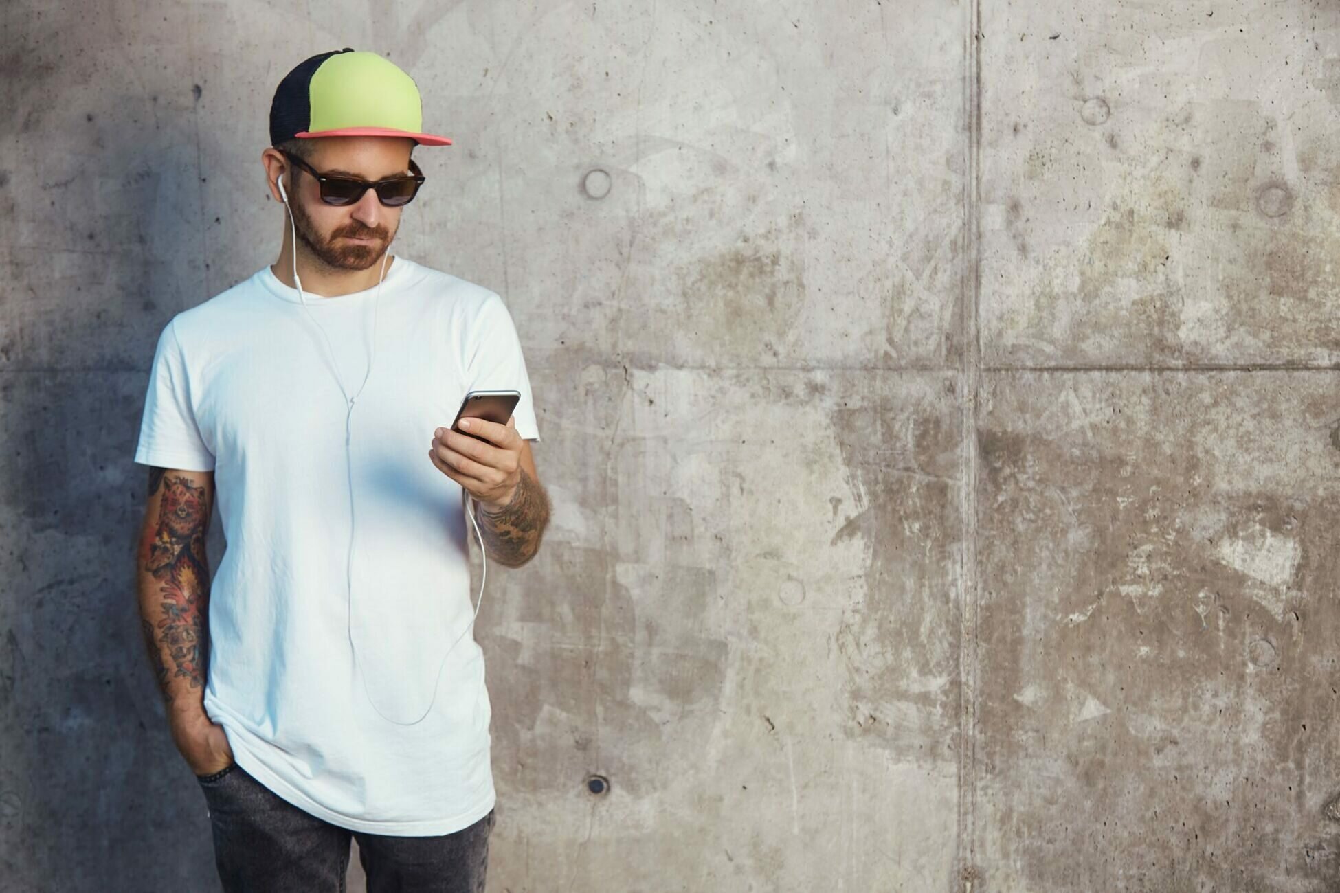 young-man-in-baseball-cap-sunglasses-and-white-blank-t-shirt-reading-something-on-his-smartphone-standing-next-to-a-gray-concrete-wall